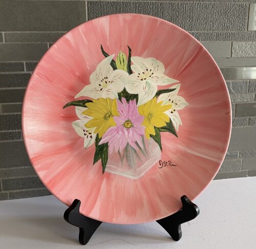 Photo of a pink hand painted plate with white lilies and pink and yellow daisies in a glass vase