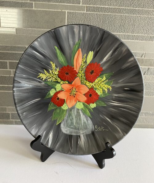 Photo of a black hand painted plate with mixed orange and red flowers in a clear glass vase